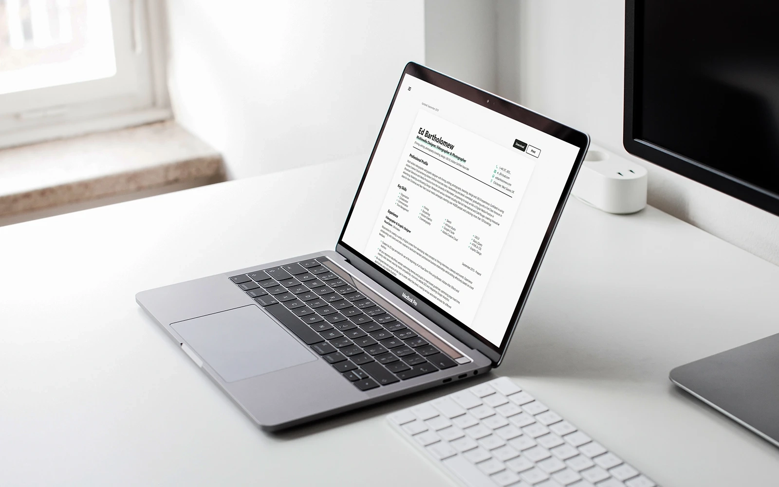 A laptop in a clean and modern room on a white desk displaying the CV page of the Ed Bartholomew website