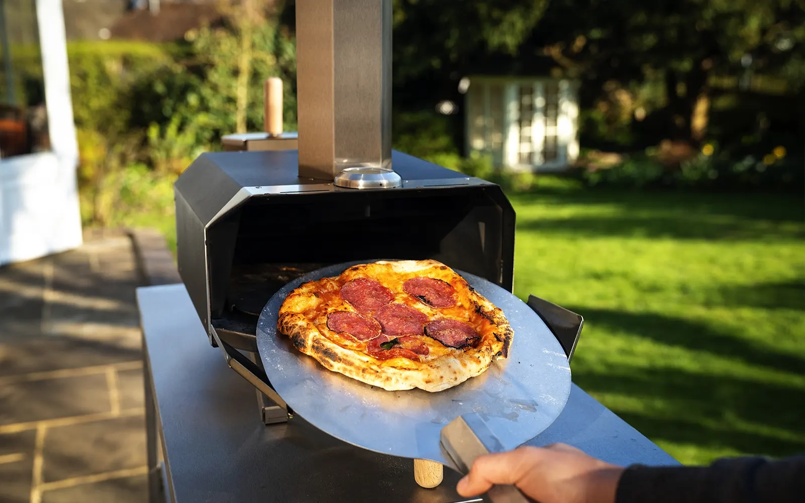 Close-up of a cooked pizza being taken out of a pizza oven on a pizza peel in a garden