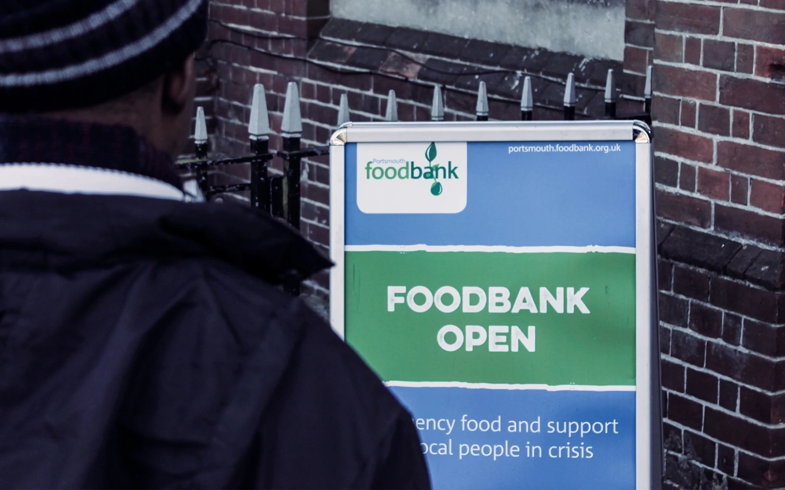 A gritty still from the Portsmouth Food Bank short documentary, taken over the shoulder of a food bank user staring at the foodbank ‘open’ sign