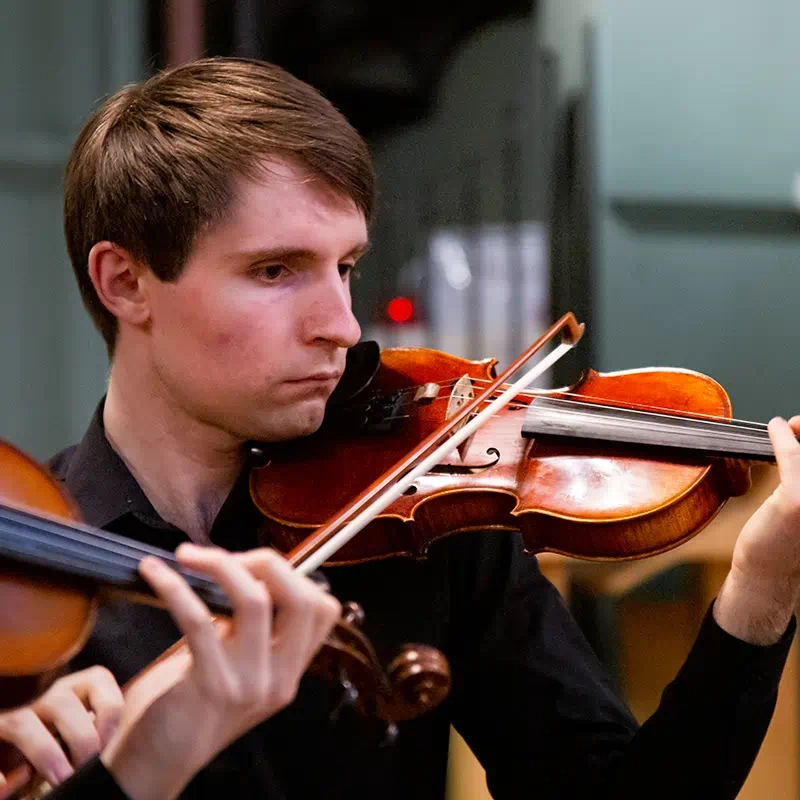 A close-up photograph of a young man playing a violin in a concert taken for use as promotional material by the Queen Mary Music Society