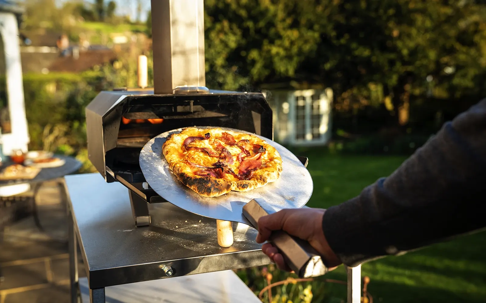 Close-up of a cooked pizza being taken out of a pizza oven on a pizza peel in a garden